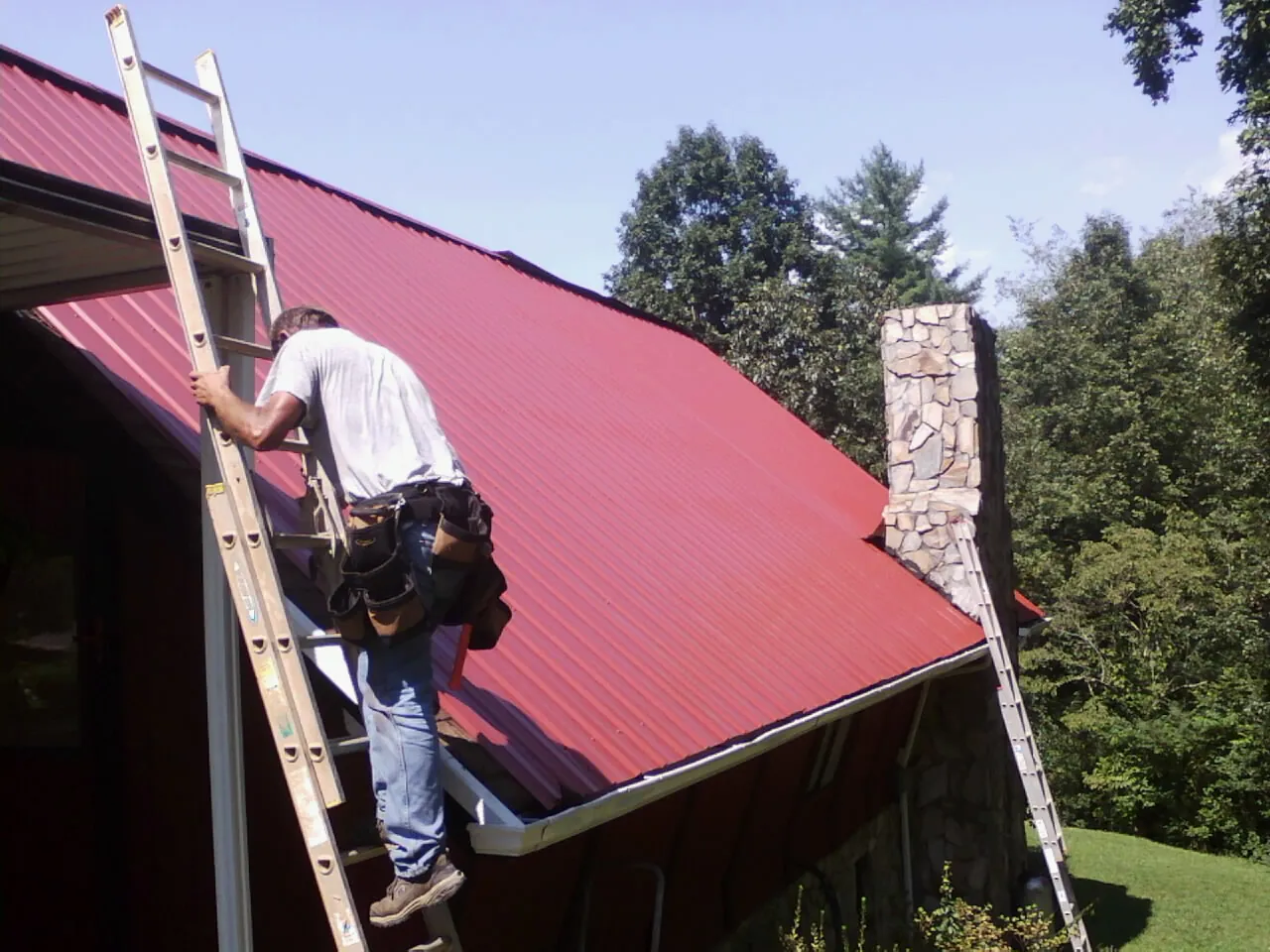 Roofer on a ladder beside a red metal roof and stone chimney — Soffit & Fascia Repair by Roofing Brothers in Somerville